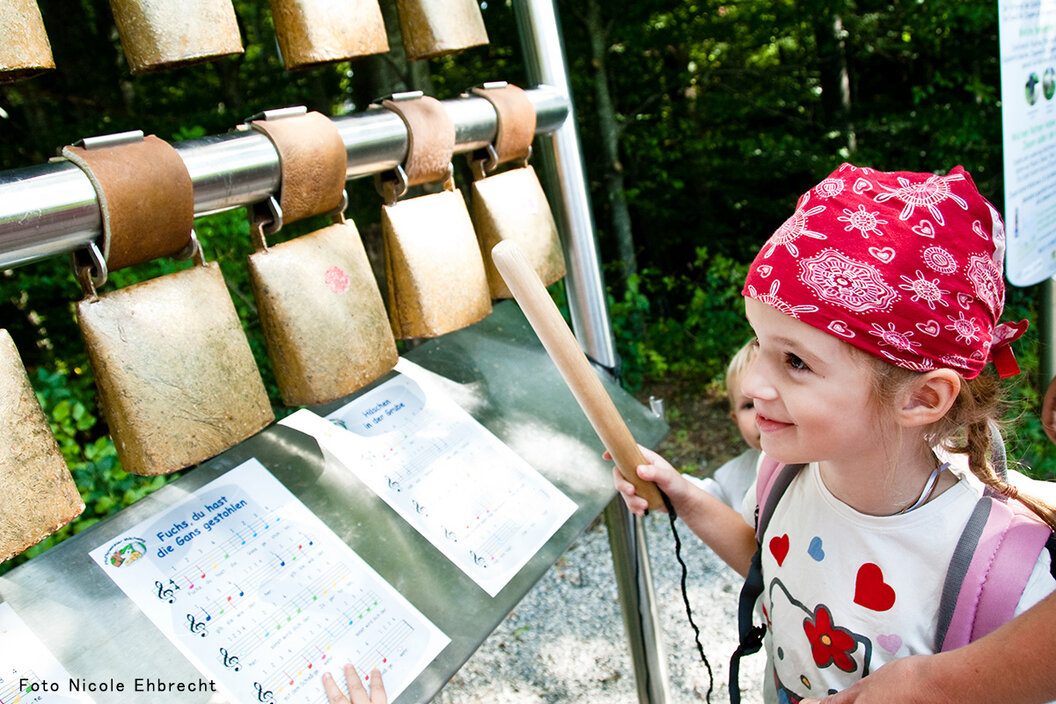 kleines Mädchen das mit einem Stock an einem Glockenspiel mit Kuhglocken beim Pfaffenwinkler Milchweg spielt