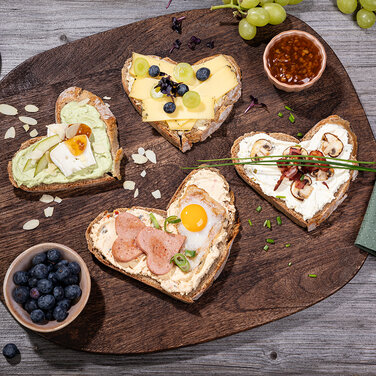leckere Brotzeit, in Herzchen form geschnittenes Brot mit verschiedenen Aufschnitten und Aufstrichen serviert auf einem rustikalen Holzbrett