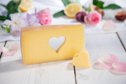 cremiges, feines Käsestück mit einem Herz ausgestochen auf einem weißen Brett mit Blüten verziert