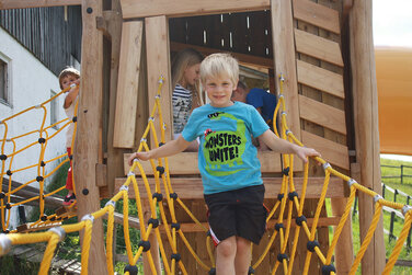 Kinder balancieren über eine Strickleiter auf dem wunderbaren Spielplatz hinter einer urigen Almhütte mit Heumilchspezialitäten